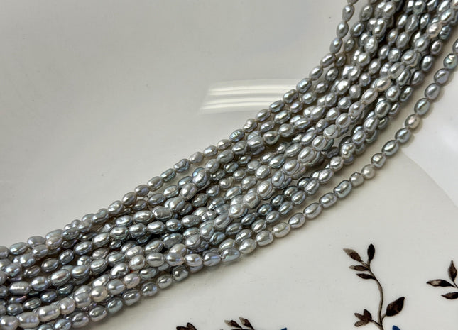 May include: Strands of gray freshwater pearls. The pearls are oval-shaped and have a lustrous sheen. The pearls are displayed on a white plate with a floral design.
